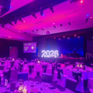 Ballroom setup for a formal event with purple lighting, round tables in white cloths and black sashes, and a stage featuring glowing '2026' letters with screens reading 'Formal 2026'.