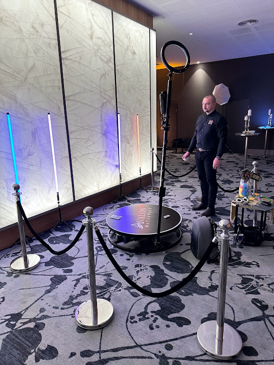 Sbollard | Osmic Productions Staff Member Stands Beside a Circular Photo Booth with a Ring Light in a Showroom, with Led Light Tubes and Rope Barriers Nearby.