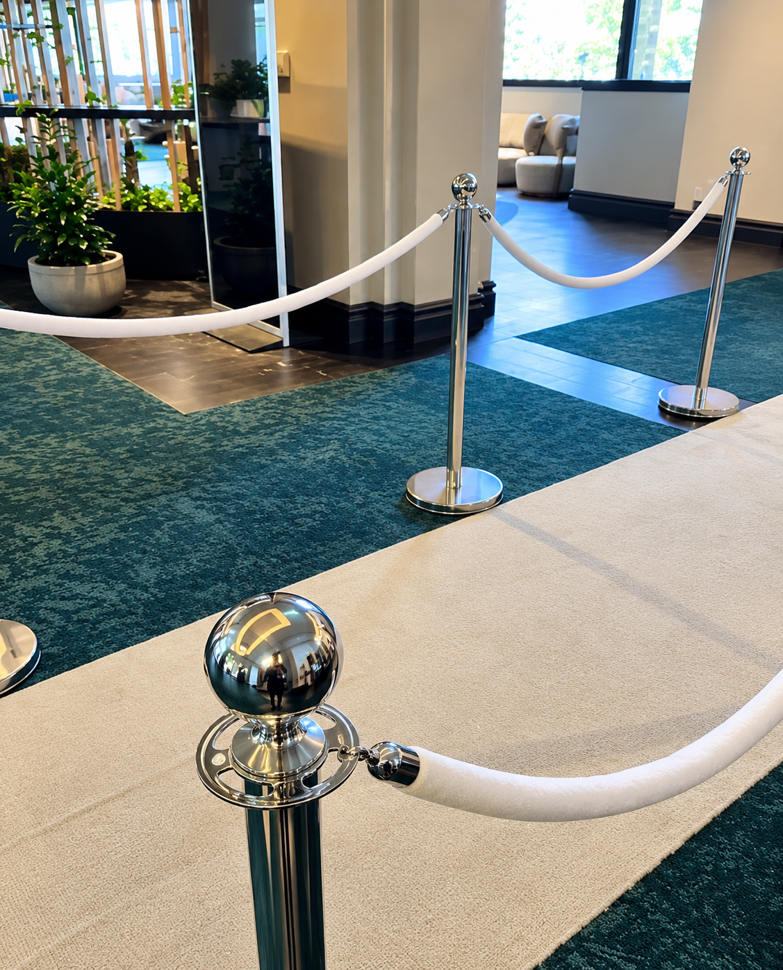 Sbollards | Osmic Productions Queue Area with Chrome Stanchions and White Ropes in a Modern Lobby, Teal Carpet and Seating Area in Background