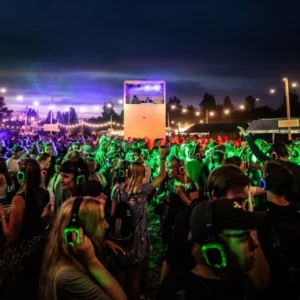 Crowd at an outdoor night festival wearing glowing headphones, with a central elevated DJ booth and string lights overhead.