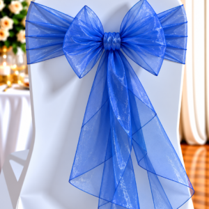 White chair decorated with a large blue organza bow for a formal event or wedding reception set-up, blurred floral background behind.