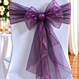 Organza Plum | Osmic Productions White Chair Draped in Fabric with a Large Translucent Purple Bow Tied at the Back, for a Formal Event Far Background Hints Candles and Flowers