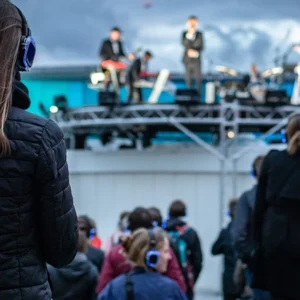 Person in a black jacket wearing blue headphones watching a live band on an outdoor stage.