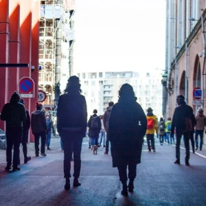Silhouetted pedestrians walking away down a narrow city street framed by brick buildings; a small dog is in the center.