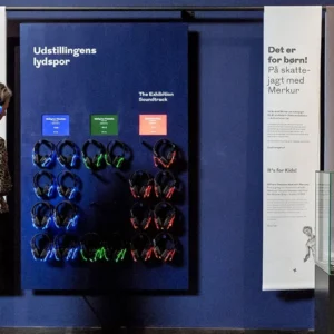 Woman in a leopard print jacket wears blue headphones, examining an exhibit panel about soundtracks at a museum display.