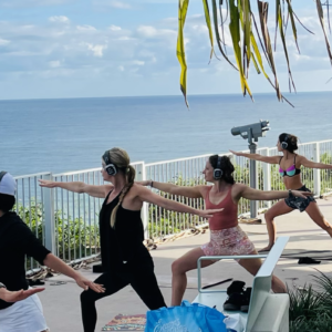 Group of people practicing yoga on a sunlit terrace by the ocean, wearing over-ear headphones and facing the water in sun-drenched poses.