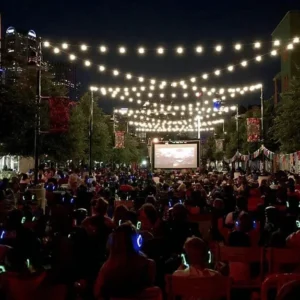Outdoor nighttime street cinema with a large screen, string lights overhead, and a crowd wearing glowing wristbands.