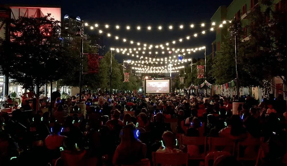 Silentcinemaoutdoorbsdmobilewebp | Osmic Productions Outdoor Nighttime Street Cinema with a Large Screen, String Lights Overhead, and a Crowd Wearing Glowing Wristbands.