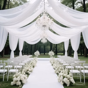 Outdoor wedding ceremony setup with white chairs along a white aisle, lined with white flowers and draped fabric, under crystal chandeliers at a park.