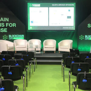 Conference stage setup for a Blockchain Expo panel with four white chairs and a glass table, a large screen showing sponsor logos, and green lighting.